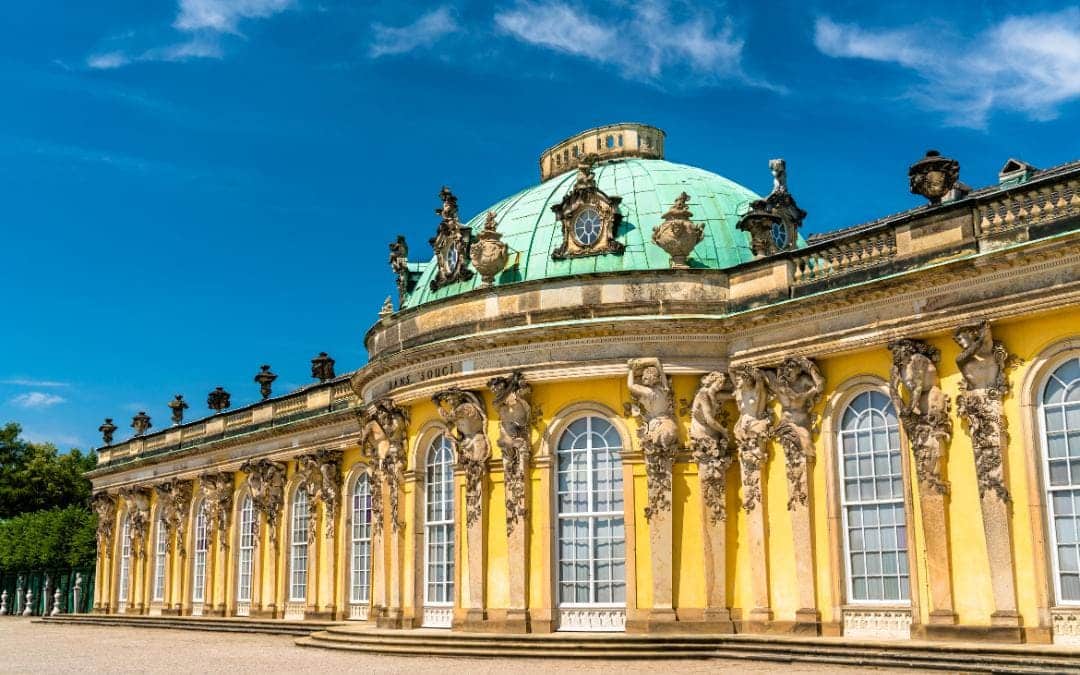 Schloss Sansscouci, Potsdam - Schrägansicht der barocken gelben Schlossfassade mit hohen Sprossenfenstern, Kapitellen mit Figuren, sowie grüner Kuppel - angiestravelroutes.com