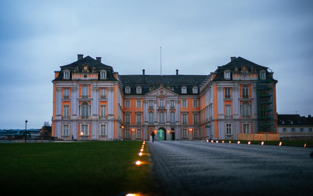 Schloss Augustusburg, Brühl, Nordrhein-Westfalen – Fassade des Schlosses von der beleuchteten Zufahrtsstraße aus kurz vor Einbruch der Dunkelheit fotografiert