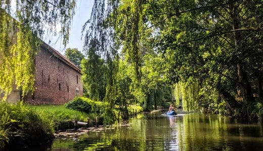 Spreewald, Brandenburg - Weeping willow and other trees over a watercourse, on the left in the picture the wall of a brick house, on the watercourse a paddler in his canoe - angiestravelroutes.com