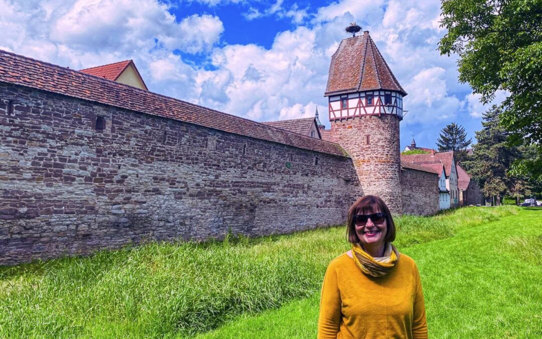 Weil der Stadt, Baden-Württemberg: Angelika Klein with sunglasses and yellow sweater on the meadow in front of the city wall and the stork tower, on the stork tower is a stork in the nest - angiestravelroutes.com