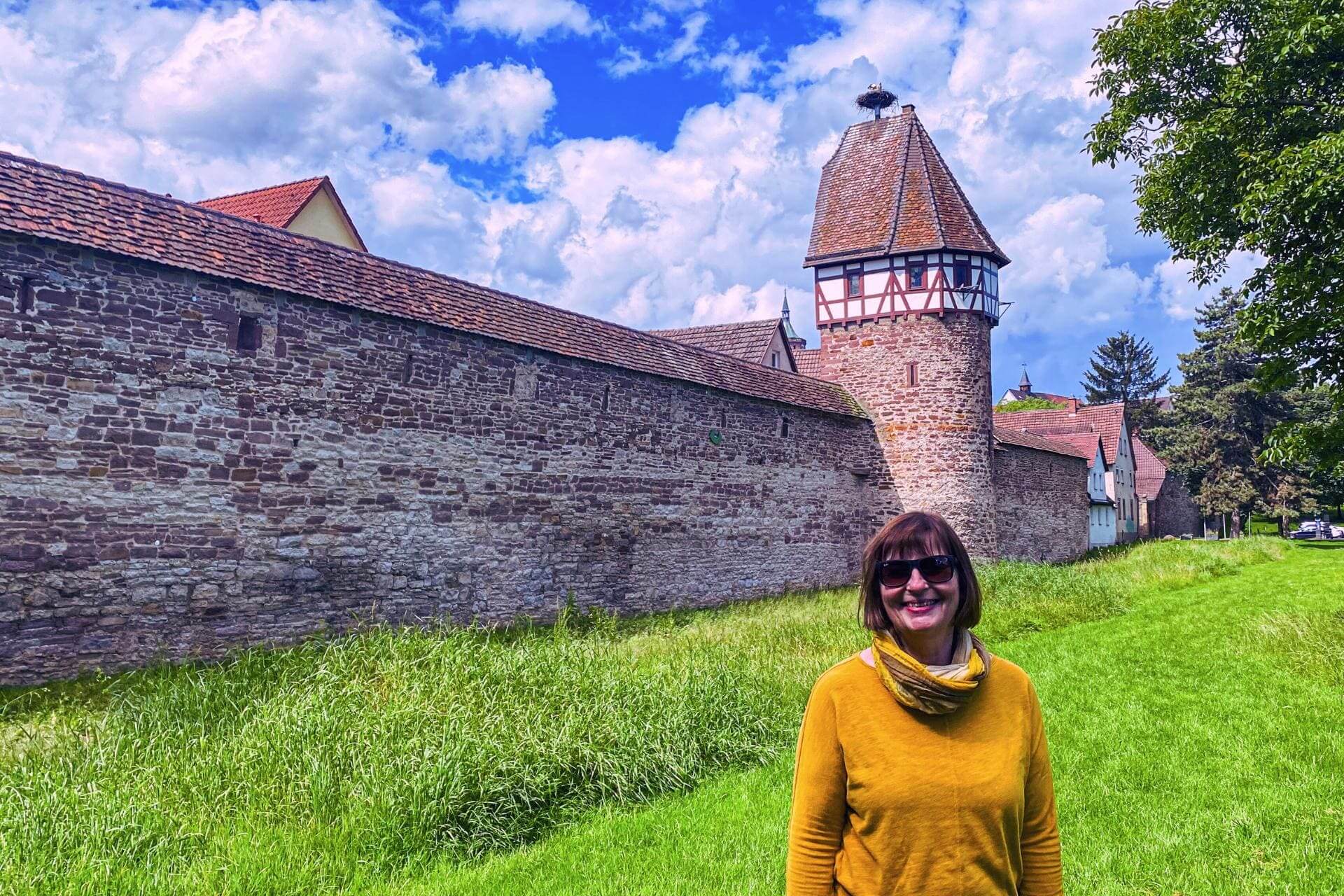 Weil der Stadt, Baden-Württemberg: Angelika Klein with sunglasses and yellow sweater on the meadow in front of the city wall and the stork tower, on the stork tower is a stork in the nest - angiestravelroutes.com