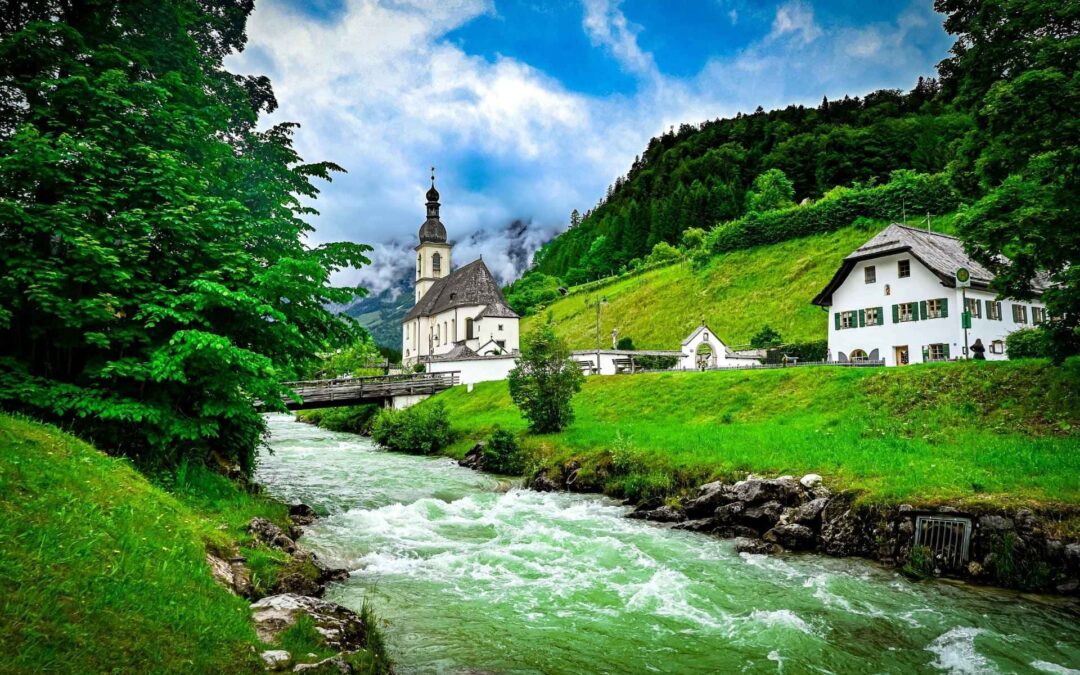 Malerwinkel Ramsau, Berchtesgadener Land: The church of St. Sebastian stands picturesquely behind the wooden bridge over the Ramsauer Ache. On the right is a typical Bavarian house with green shutters and a gabled roof - angiestravelroutes.com