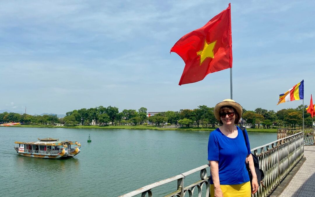 Vietnam, Hué, Truong-Tien-Brücke am Parfüm-Fluss: Frau mit Sonnenbrille, Sonnenhut, königsblauem T-Shirt und gelbem Rock steht unter der vietnamesischen Flagge auf der Brücke (gelber Stern auf rotem Grund), dahinter das baumbestandene Nordufer, links im Hintergrund ein Boot auf dem Parfüm-Fluss - angiestravelroutes.com