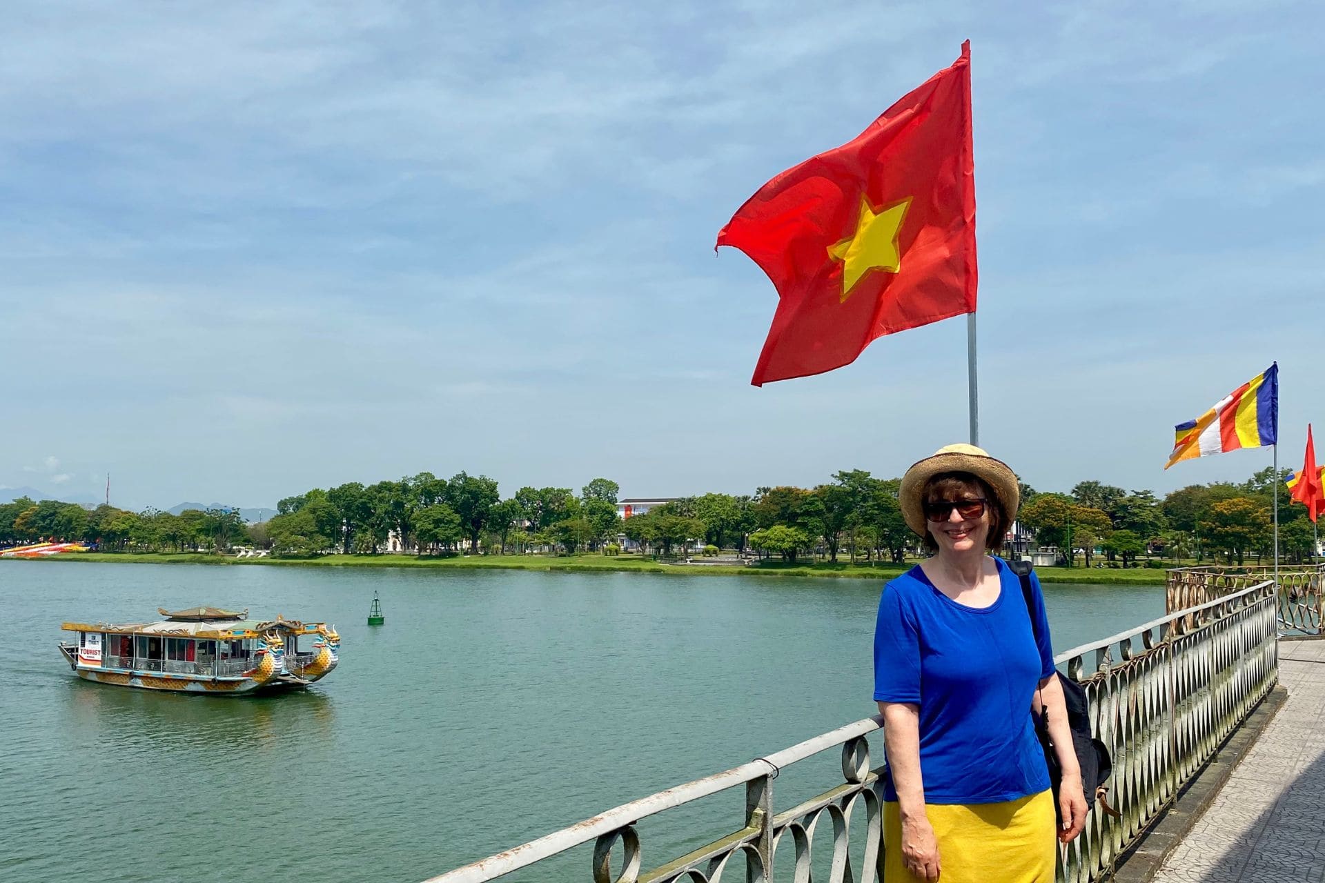 bb-morue-mai-2025-vietnam-hue-angelika Vietnam, Hué, Truong-Tien-Brücke am Parfüm-Fluss: Frau mit Sonnenbrille, Sonnenhut, königsblauem T-Shirt und gelbem Rock steht unter der vietnamesischen Flagge auf der Brücke (gelber Stern auf rotem Grund), dahinter das baumbestandene Nordufer, links im Hintergrund ein Boot auf dem Parfüm-Fluss - angiestravelroutes.com