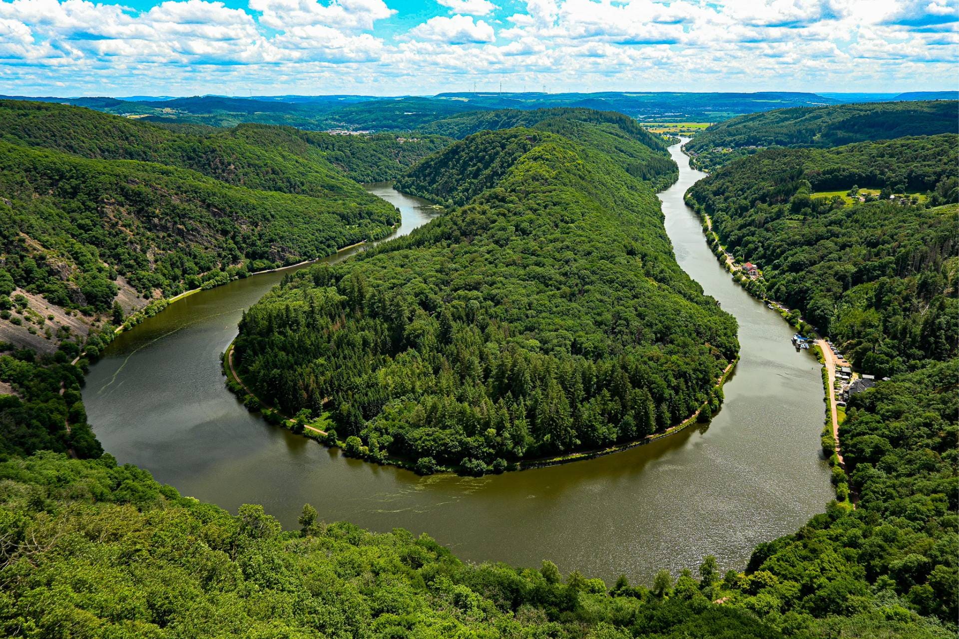 bb-saarland-attractions-Saar Loop The Saar Loop near Mettlach — A wide river winds its way beneath a partly cloudy sky through dense, green forested hills, forming a striking bend in the landscape.