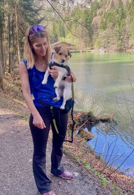 Berglsteiner See, Trol, Österreich - junge Frau in Jeans und königsblauem Top mit kleinem braun-weißem Hund auf dem Arm am Seeufer - angiestravelroutes.com