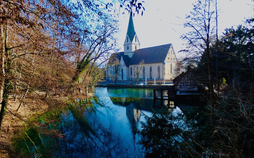 Blautopf, Blaubeuren: Die tiefblaue Karstquelle im Winter vor der Klosterkirche, eingerahmt von Bäumen mit verfärbtem Laub - angiestravelroutes.com