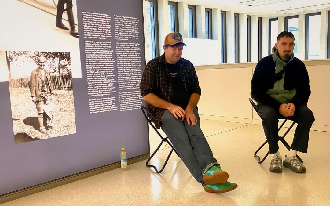 NS-Dokumentationszentrum München - Christoph Zahner (links im Bild) mit gelber Baseball-Cap, kariertem Hemd, Jeans und grünen Turnschuhen auf einem Klappstuhl sitzend, neben einer bebilderten und beschrifteten Wand. Rechts von ihm, ebenfalls auf einem Klappstuhl, ein schwarz gekleideter junger Mann mit Oberlippen- und Kinnbart - angiestravelroutes.com