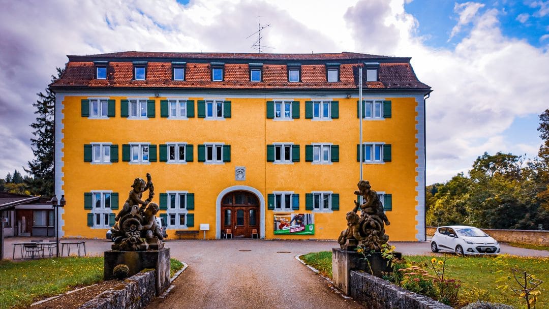 Grafeneck Castle, front view of the Renaissance castle, yellow-painted façade, green shutters, with groups of figures to the right and left of the driveway - angiestravelroutes.com