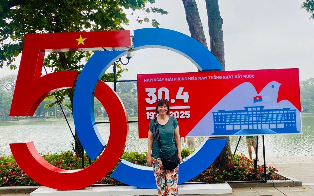 Fotospot am Hoan-Kiem-See, Hanoi: Frau mit moosgrünem T-Shirt und bunter Hose steht for dem 50-Jahr-Feier-Schild in rot-weiß-blau, im Hintergrund der See. - angiestravelroutes.com