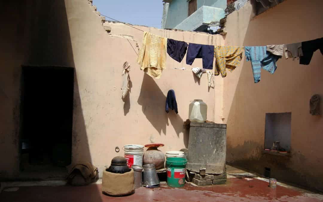 Patio in an Indian house in the countryside - a washing line with clothes is stretched across the corner between two walls, on the floor is a tin water canister, several buckets made of plastic and tin, the walls are painted yellowish-pink, the floor rust-red - angiestravelroutes.com