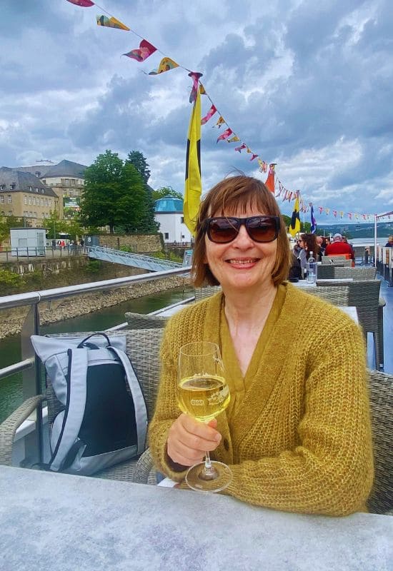 Angelika Klein on the "Wein on the Water" Rhine cruise, holding a glass of white wine, with the Koblenz Water Level House in the background