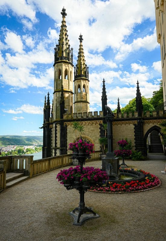The towers of the chapel at Stolzenfels Castle, with the Rhine in the background and the castle courtyard, featuring a flower bed and a fountain, in the foreground
