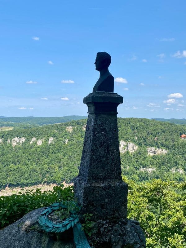 Hauff-Denkmal bei Schloss Lichtenstein: Büste des Dichters auf einem Sockel an der Kante des Albtraufs vor strahlendblauem Himmel, im Hintergrund die bewaldeten Berge der Schwäbischen Alb - angiestravelroutes.com