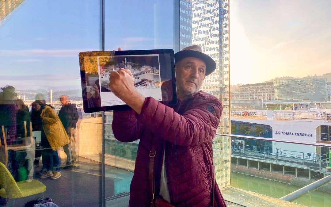 Museum Lentos, Linz - Mann mit Hut und Bart hält ein Tablet hoch, auf dem man ein altes Foto einer Industrieanlage sieht, auf das er deutet; im Hintergrund ein Fenster - angiestravelroutes.com