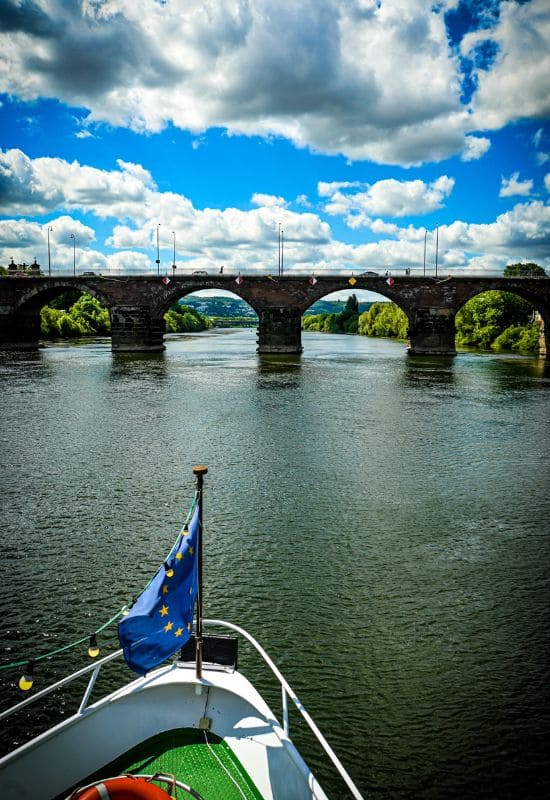 Römerbrücke mit ihren mächtigen antiken Pfeilern unter blauem Himmel mit weißen Wolken, im Vordergrund der Schiffsbug