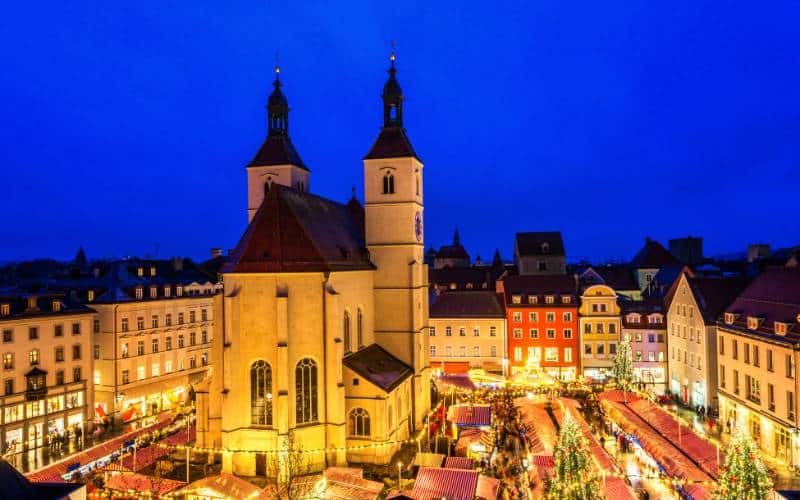 Luftaufnahme des Christkindlmarkts auf dem Regensburger Neupfarrplatz am Abend - die Neupfarrkirche ist umgeben von Weihnachtsständen, auf dem Markt stehen mehrere beleuchtete Tannenbäume. - angiestravelroutes.com