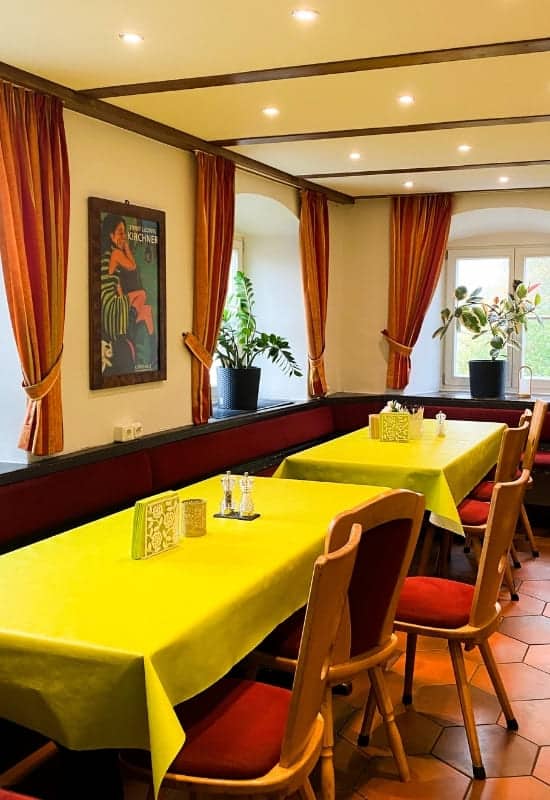 Speisesaal Schloss Aspenstein, Kochel am See - orangefarbene Vorhänge, gelbgrüne Wachstischdecken, helle Holzmöbel - angiestravelroutes.com