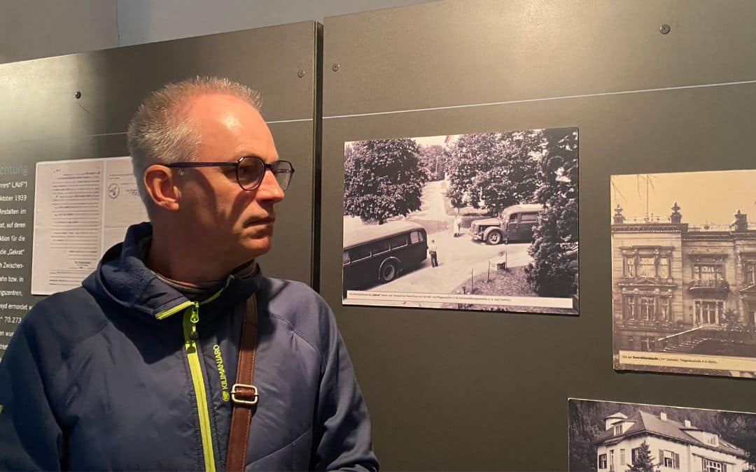 Mark Kaiser, Gedenkstätte Schloss Hartheim - ein Mann mit Brille und kurzem grauem Haar steht schräg vor einer Wand mit alten Schwarz-Weiß-Fotos - angiestravelroutes.com