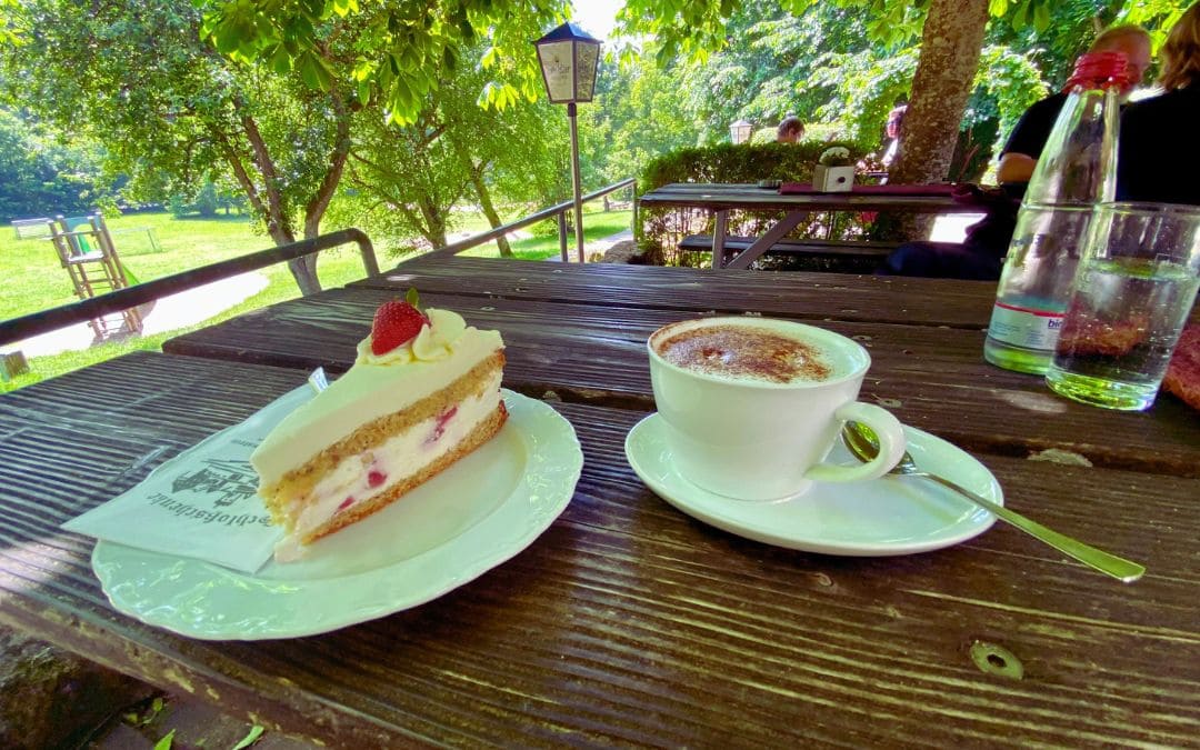 Schlossschenke: Käsesahnetorte mit Erdbeeren , Cappuccino und eine kleine Flasche Minteralwasser mit Glas auf einem Holztisch im Biergarten, im Hintergrund Laubbäume und links im Bild ein Spielplatz - angiestravelroutes.com