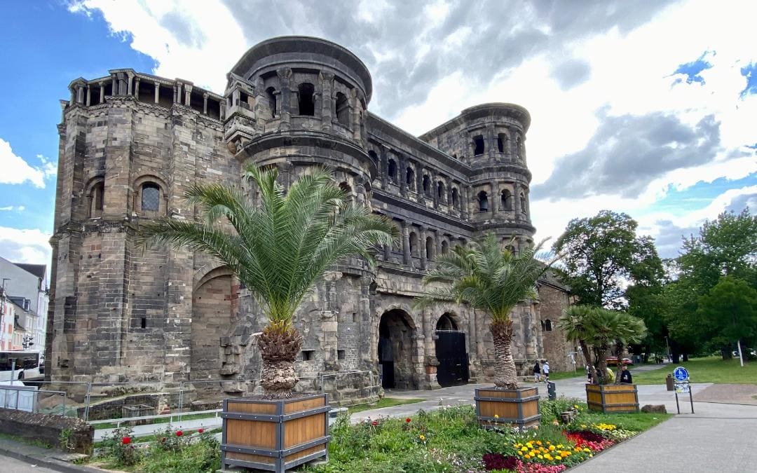 Porta Nigra mit Blumenbeeten und Palmen vor blauem Himmel mit Schönwetterwolken
