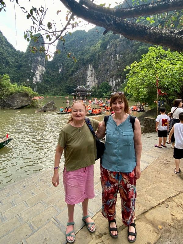 Trang An, Vietnam - zwei mittelalte Frauen am Flussufer, im Hintergrund viele Ruderboote mit Touristen auf dem Fluss, eine Pagode und die bewaldete Karstlandschaft (hier der "Lying Dragon Mountain") - angiestravelroutes.com