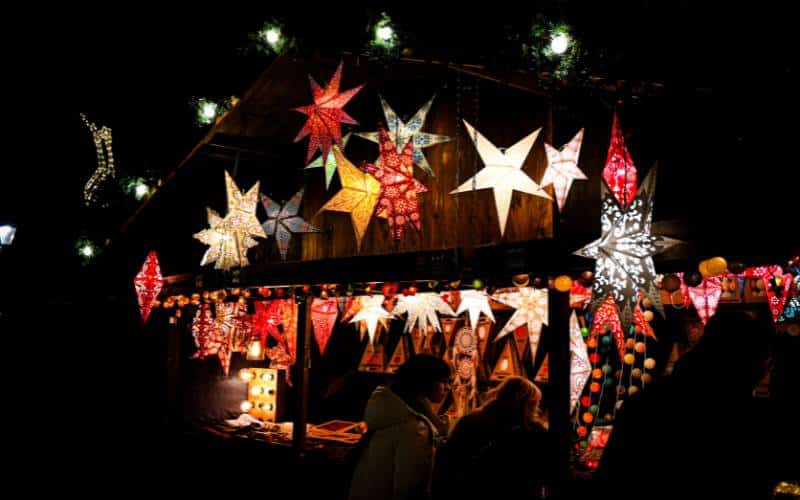 Weihnachtshütte mit leuchtenden, verzierten Papiersternen auf einem deutschen Weihnachtsmarkt am Abend. Die Hütte ist kaum sichtbar, man sieht vor allem die leuchtenden Sterne und im Vordergrund die Umrisse von zwei Frauen, die sich die Auslagen ansehen. - angiestravelroutes.com